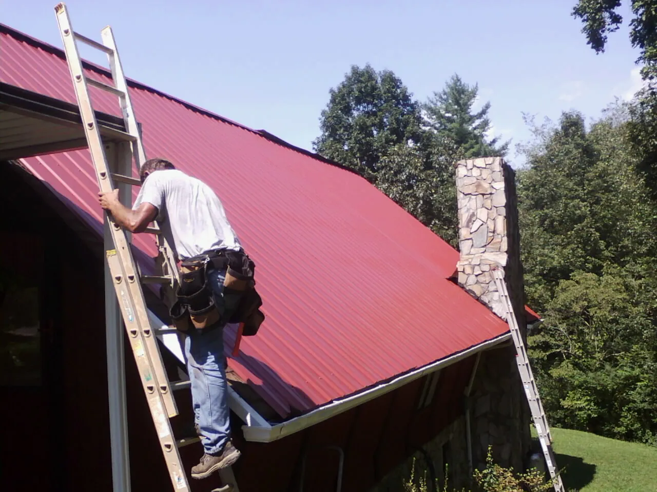 Roofer on a ladder beside a red metal roof and stone chimney — Soffit & Fascia Repair by Roofing Brothers in Kinzers